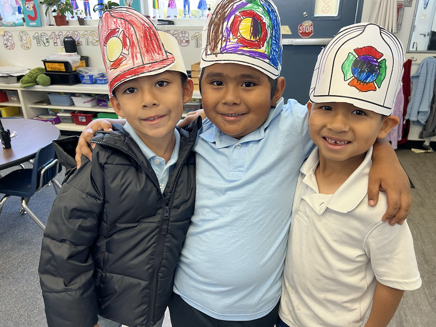 three kindergarten boys with arts around each other, wearing paper firefighter hats