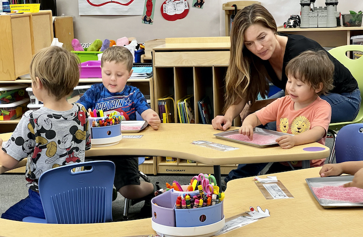 teacher leaning in with young student at table, other children at table