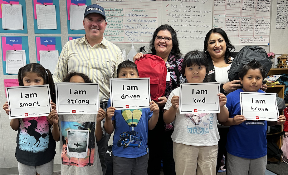 group photo of adult man and two adult women holding backpacks with five students holding signs that say I am smart, strong, driven, kind, brave