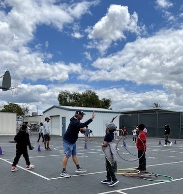 coach high-fives students while other students on blacktop with hoops and balls under bright blue sky