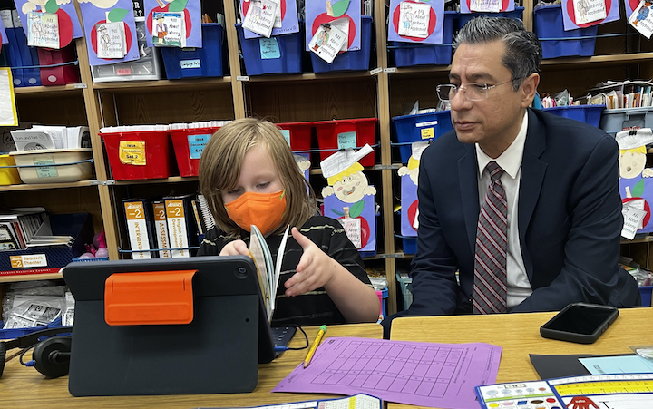 niño con máscara, trabajando con un libro y un iPad sentado junto a un hombre en la mesa del aula