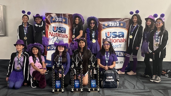 12 girls in dance team jackets purple hats posing with three trophies