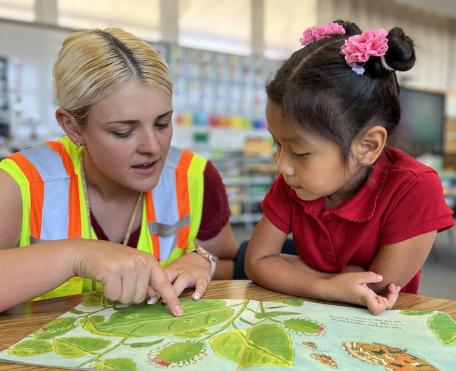 blonded woman teacher's aide reading book with girl student in red shirt