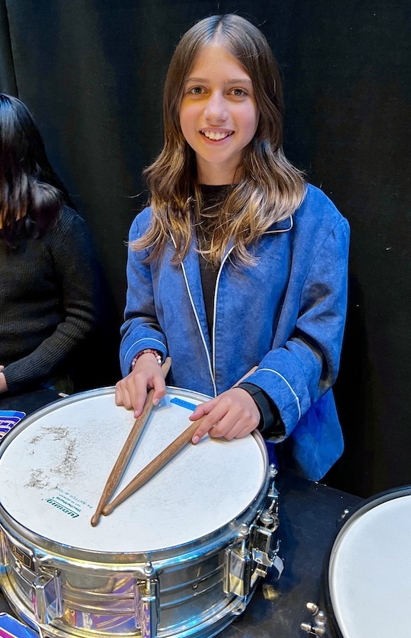 girl in blue jacket in front of drum with drumsticks
