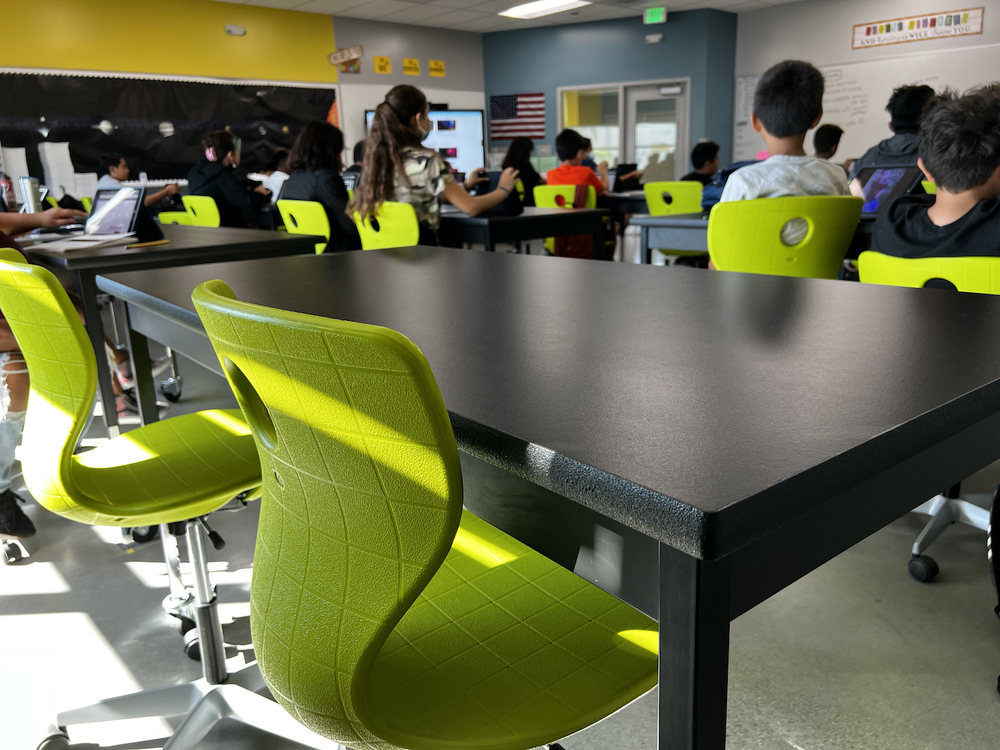 vista del aula con mesas negras, sillas verdes, espaldas de los estudiantes