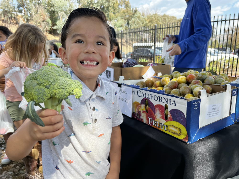 boy smiling holding up broccoli outdoors at farmers market
