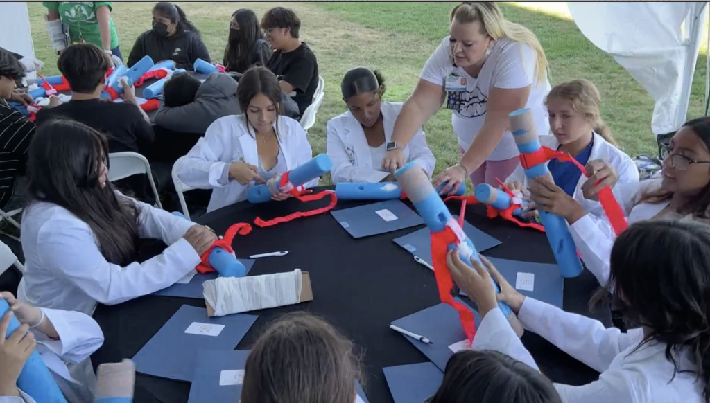 students in white lab coats around table using pool noodles to practice tourniquets