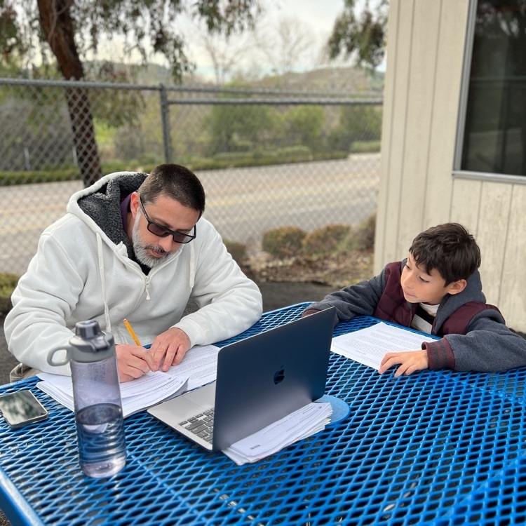 teacher with laptop listening to student read at outdoor table