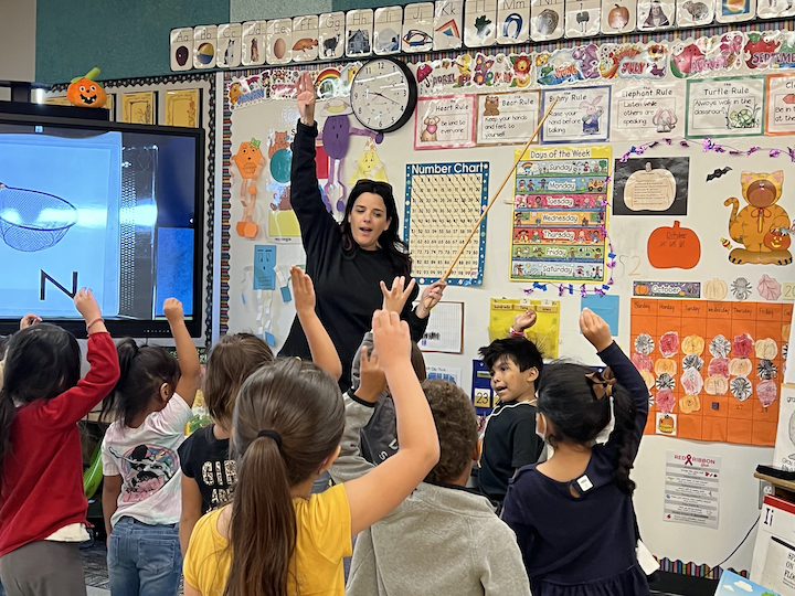 children standing in classroom raising hands facing singing teacher