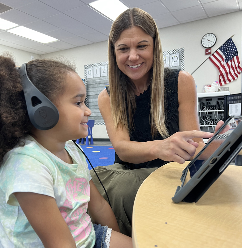 teacher pointing to ipad with young girl student wearing headphones