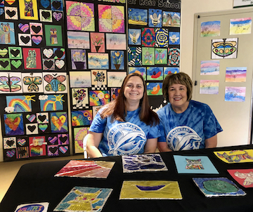 two women sitting at table surrounded by student artwork