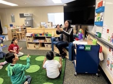 preschool teacher sitting on chair talking to children sitting on green rug